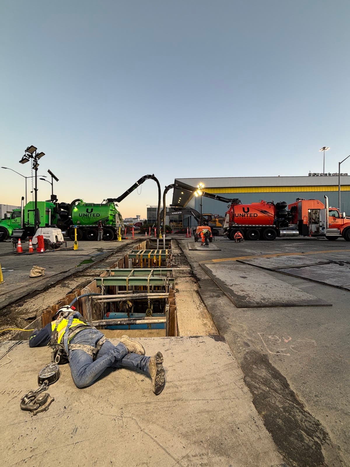 Hydrovac operator in hi-vis gear kneeling at the edge of a shored excavation, with green and orange vacuum trucks parked behind