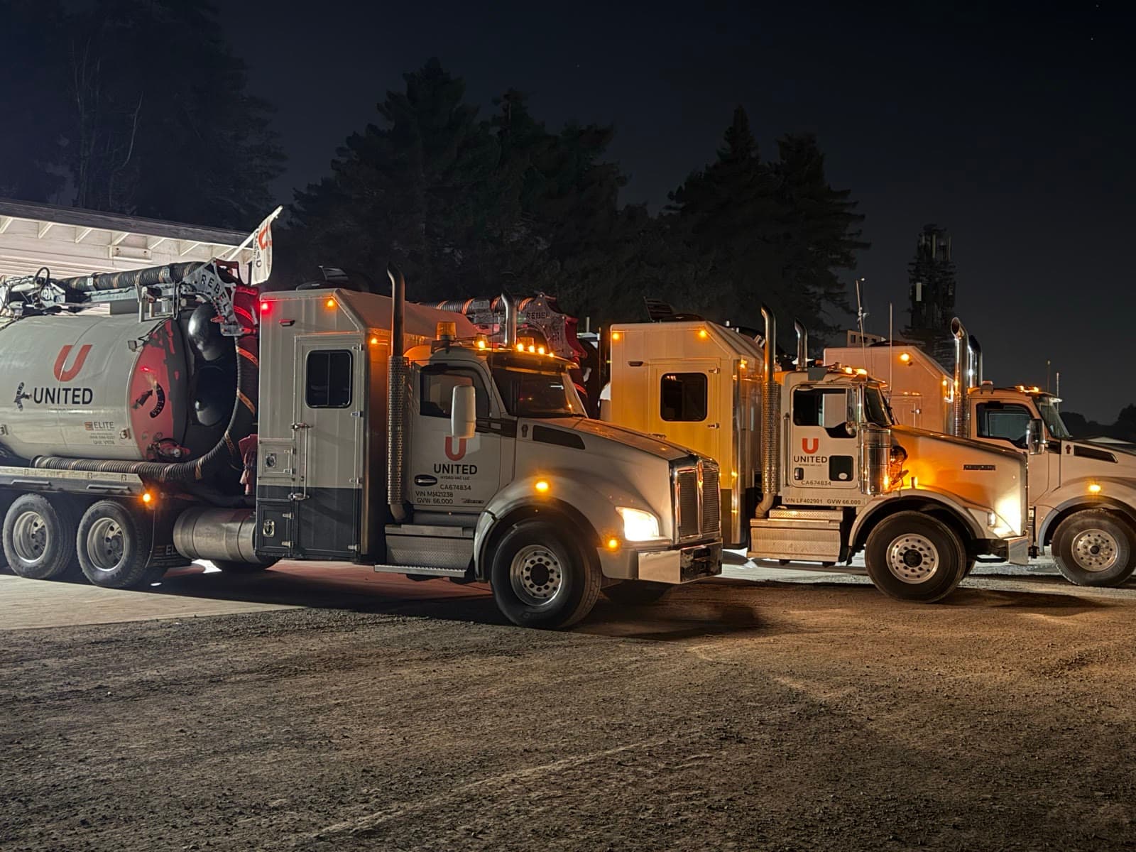 Four United Hydrovac trucks lined up side by side at night, headlights and amber marker lights on